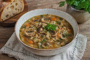 Глубокая тарелка наваристого грибного супа с перловкой, картофелем и зеленью, поданная с ломтиком хлеба