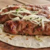 Румяные мясные люля-кебабы на тарелке с маринованным красным луком, зеленью и тонкими лепешками лаваша