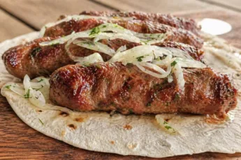 Румяные мясные люля-кебабы на тарелке с маринованным красным луком, зеленью и тонкими лепешками лаваша