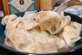 Глубокая фарфоровая миска с горкой круглых горячих пельмешек, политых желтым растопленным маслом и присыпанных свежим черным перцем
