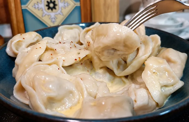 Глубокая фарфоровая миска с горкой круглых горячих пельмешек, политых желтым растопленным маслом и присыпанных свежим черным перцем