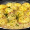 Аппетитные кусочки золотистого картофеля в густом белом сырном соусе с кусочками бекона и зеленью на сковороде