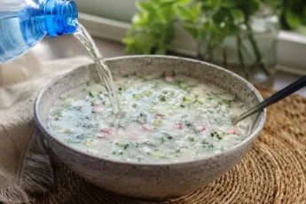 Глубокая тарелка с густой белой окрошкой, в которой видны кусочки розовой колбасы, редиса и обилие зелени