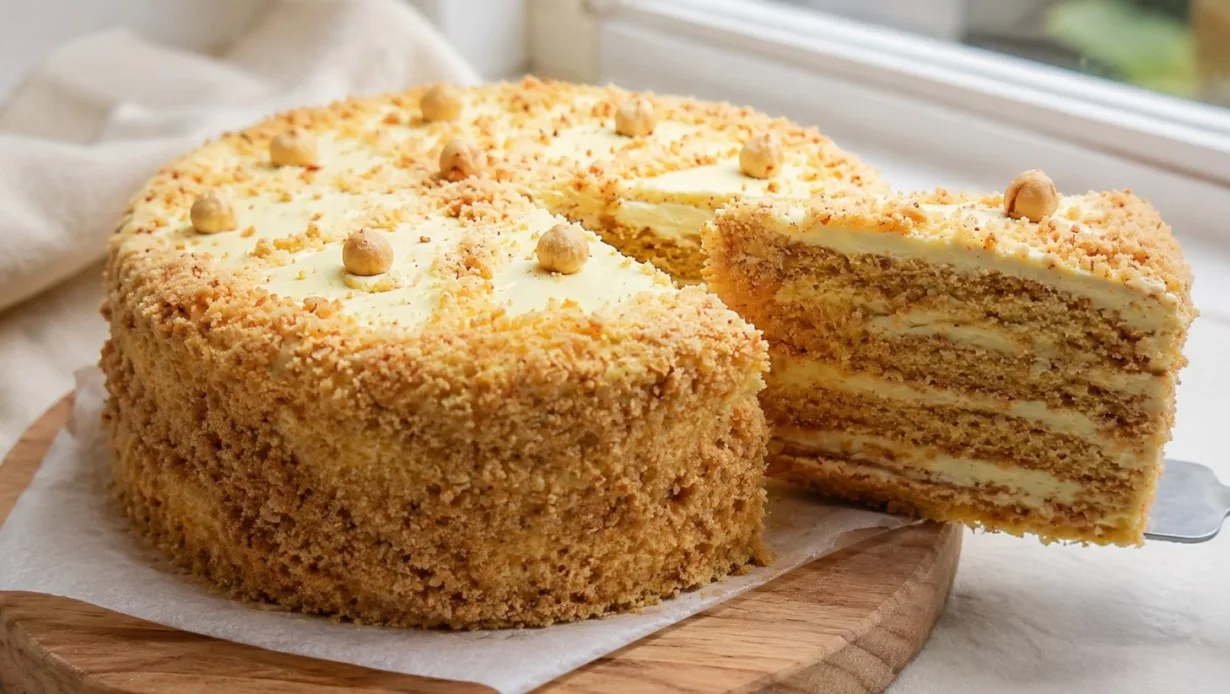 Пышный круглый торт, густо покрытый бежевым кремом и обильно обсыпанный золотистой крошкой из жареного фундука