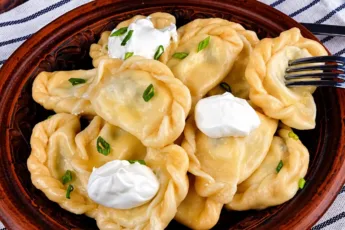 Глубокая белая миска с горячими, пухлыми свежесваренными варениками, обильно политыми желтым сливочным маслом и посыпанными колечками зеленого лука