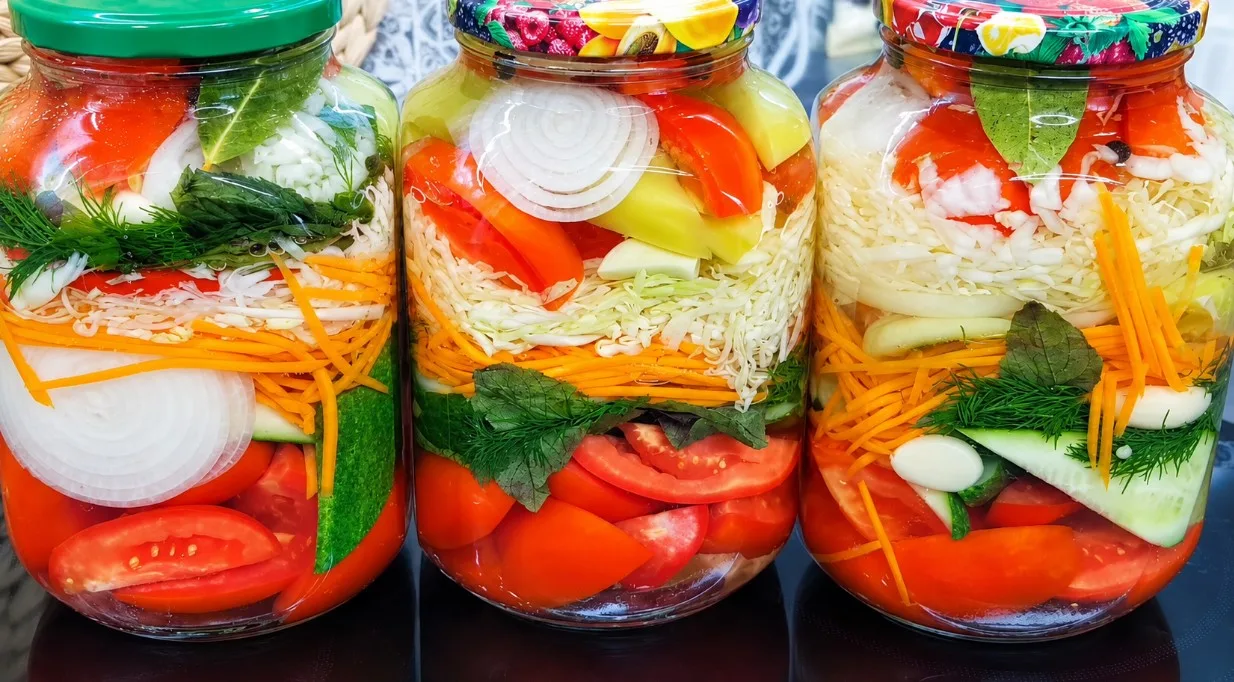 Разноцветные слои маринованных овощей с зеленью и чесноком в прозрачной стеклянных банках на зиму