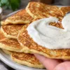 Стопка золотистых яблочных оладий на белой тарелке, политая сметанным соусом с корицей и лимоном