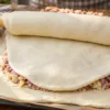 Процесс приготовления большого домашнего пирога с картошкой и фаршем на противне, верхний слой тонкого бездрожжевого теста аккуратно переносят с помощью деревянной скалки на мясную начинку