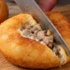 Крупный план: рука автора кухонным ножом разрезает пополам горячий пышный домашний беляш на деревянной доске, сквозь разрез видна сочная мясная начинка из фарша и пористая структура мягкого золотистого теста