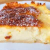 Крупный план: аппетитный порционный кусок итальянского десерта латтайоло на белой тарелке, отчетливо видна нежная кремовая текстура заварного пирога в разрезе и аппетитная румяная корочка с сахарной пудрой