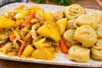 Домашние штрудли с мясом и картофелем в тарелке, крупным планом видны пышные паровые рулетики из теста с чесночной начинкой, кусочки тушеной свинины, картошка и квашеная капуста