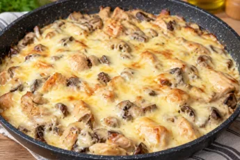 Аппетитный горячий жульен с курицей и грибами в глубокой сковороде, вся поверхность блюда покрыта плотным слоем расплавленного тягучего желтого сыра с румяной корочкой